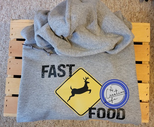 A gray hoodie with a graphic that has the words 'FAST FOOD' and an image of a deer on a yellow sign, positioned on a wooden surface.