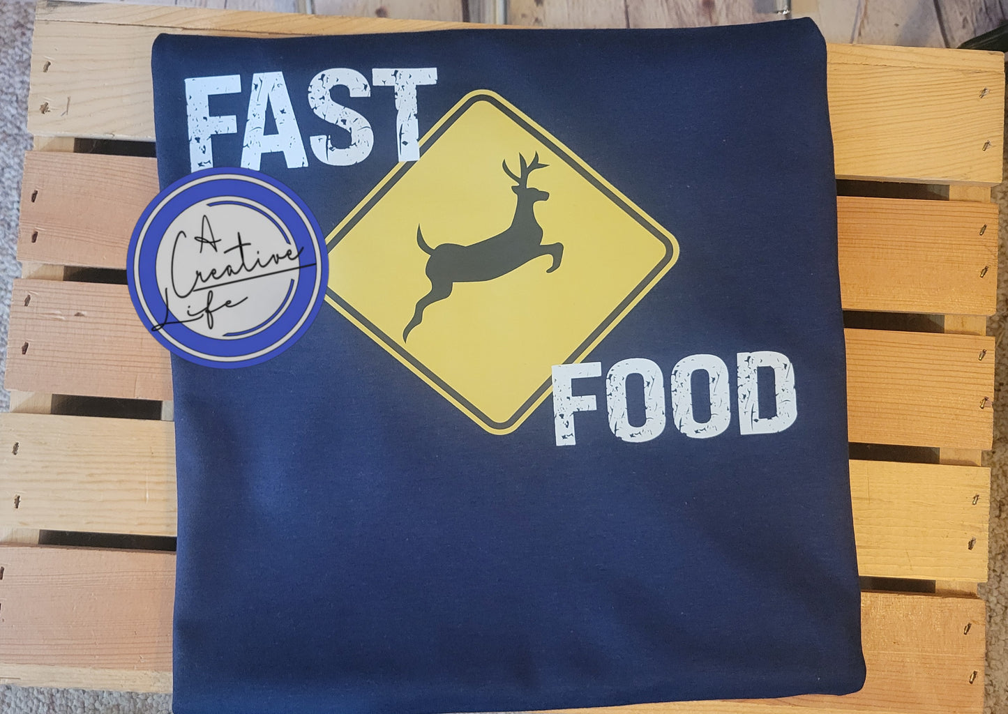 A navy blue t-shirt with a graphic that has the words 'FAST FOOD' and an image of a deer on a yellow sign, positioned on a wooden surface.