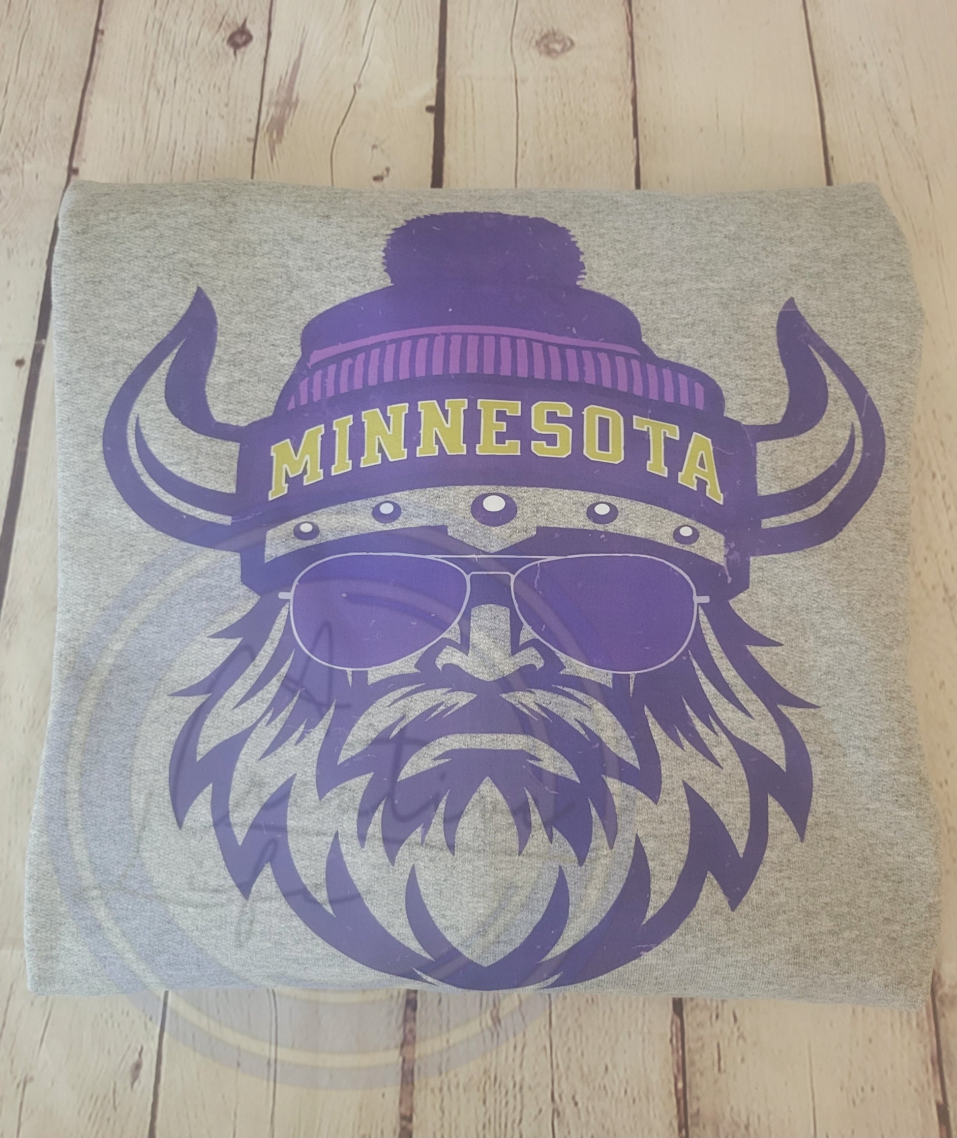 Athletic Gray Crewneck Sweatshirt with purple graphic of a Viking wearing sunglasses and a 'Minnesota' beanie on a wooden surface.